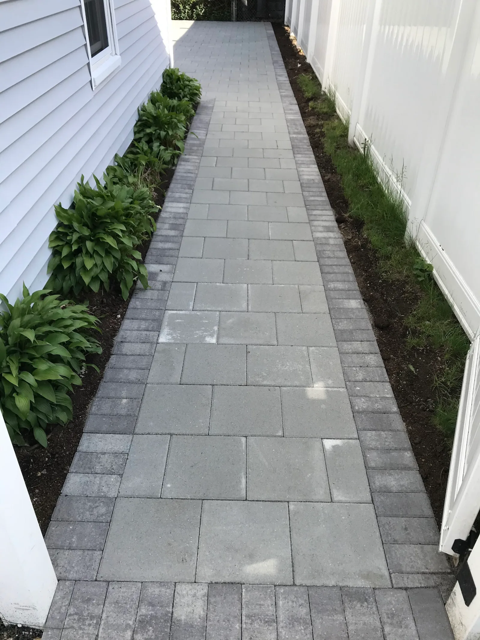 Paver walkway with a dark border running alongside a house between planting beds